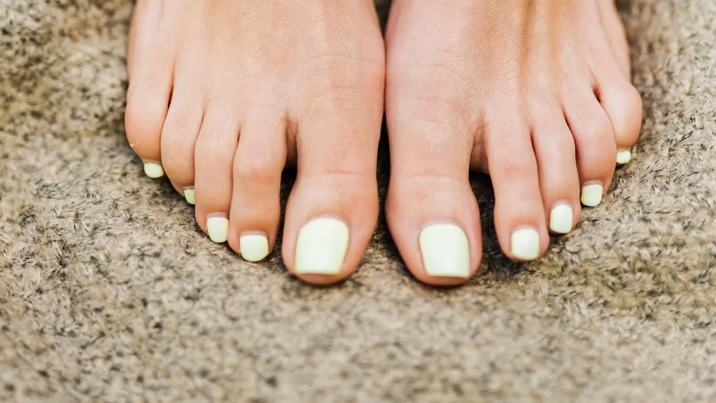 Dos pies descalzos apoyados sobre una manta beige, con las uñas pintadas de verde pálido, vistos de frente y de cerca: las uñas de los pies que se revisan cada noche