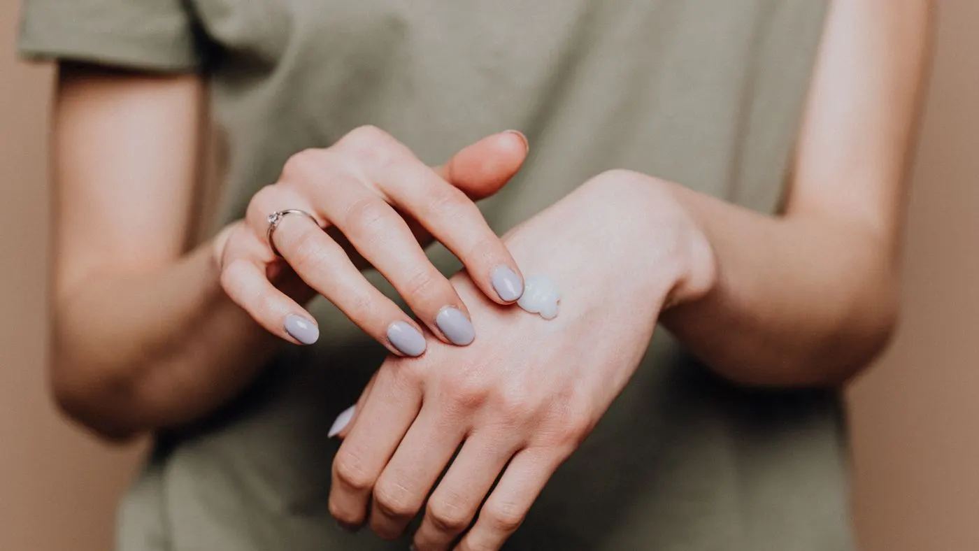 Manos de una persona que se extiende una crema blanca sobre el dorso de la mano, con una camiseta verde oliva al fondo y luz suave
